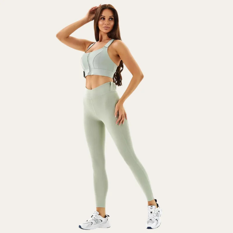 Mulher a usar o soutien desportivo Alba verde acinzentado com leggings a condizer e ténis brancos, com a mão no cabelo